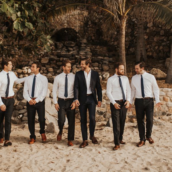 伴郎和新郎在沙滩前的老石头结构和棕榈树“class=