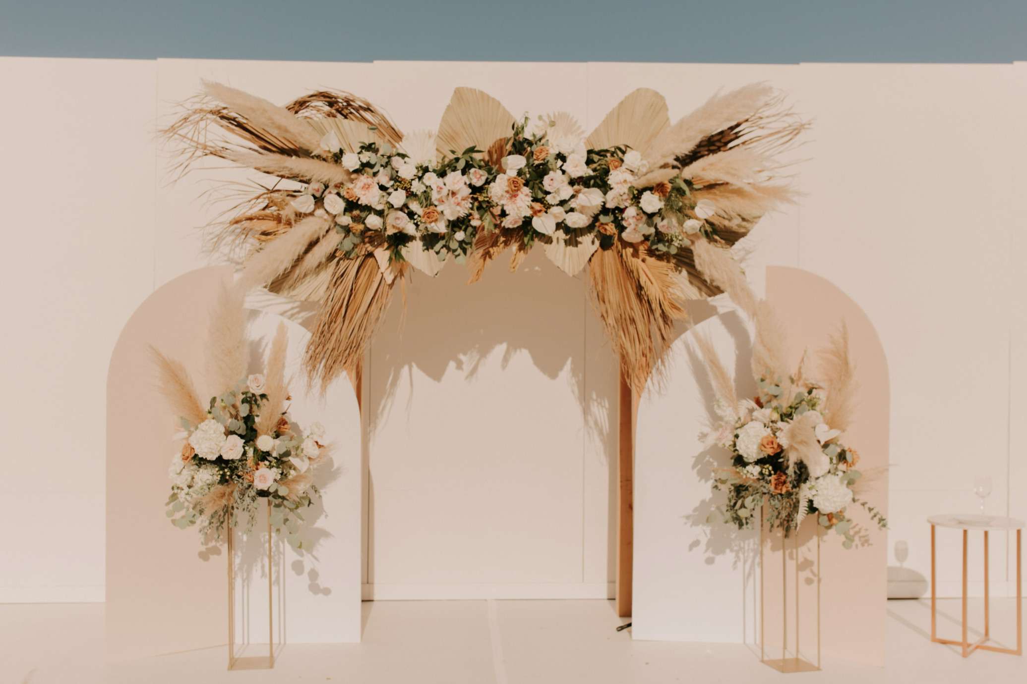 婚礼拱门装饰着白色，粉红色和金色的花朵，干棕榈叶和潘帕斯草
