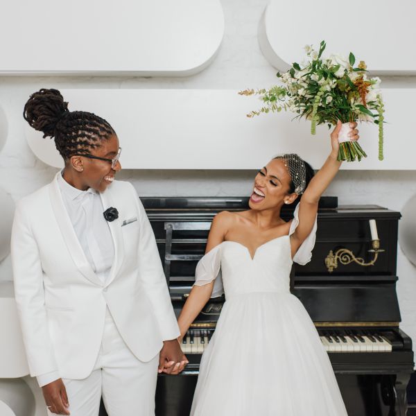 两位新娘手牵着手，在钢琴前摆姿势，左边的新娘举起她的花束，微笑着