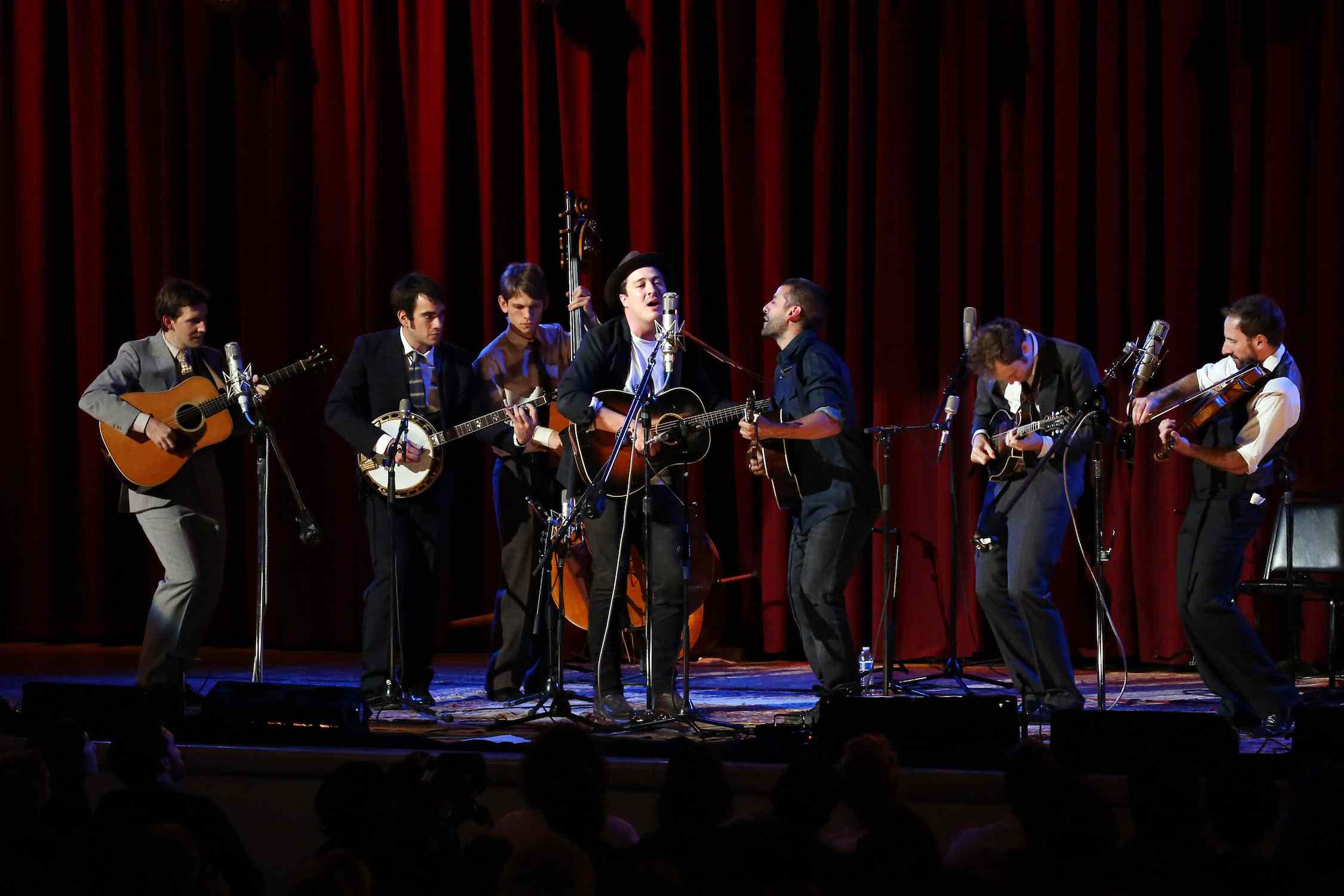 马库斯·芒福德和奥斯卡·艾萨克在台上共用一个麦克风，一群音乐家在他们身后演奏