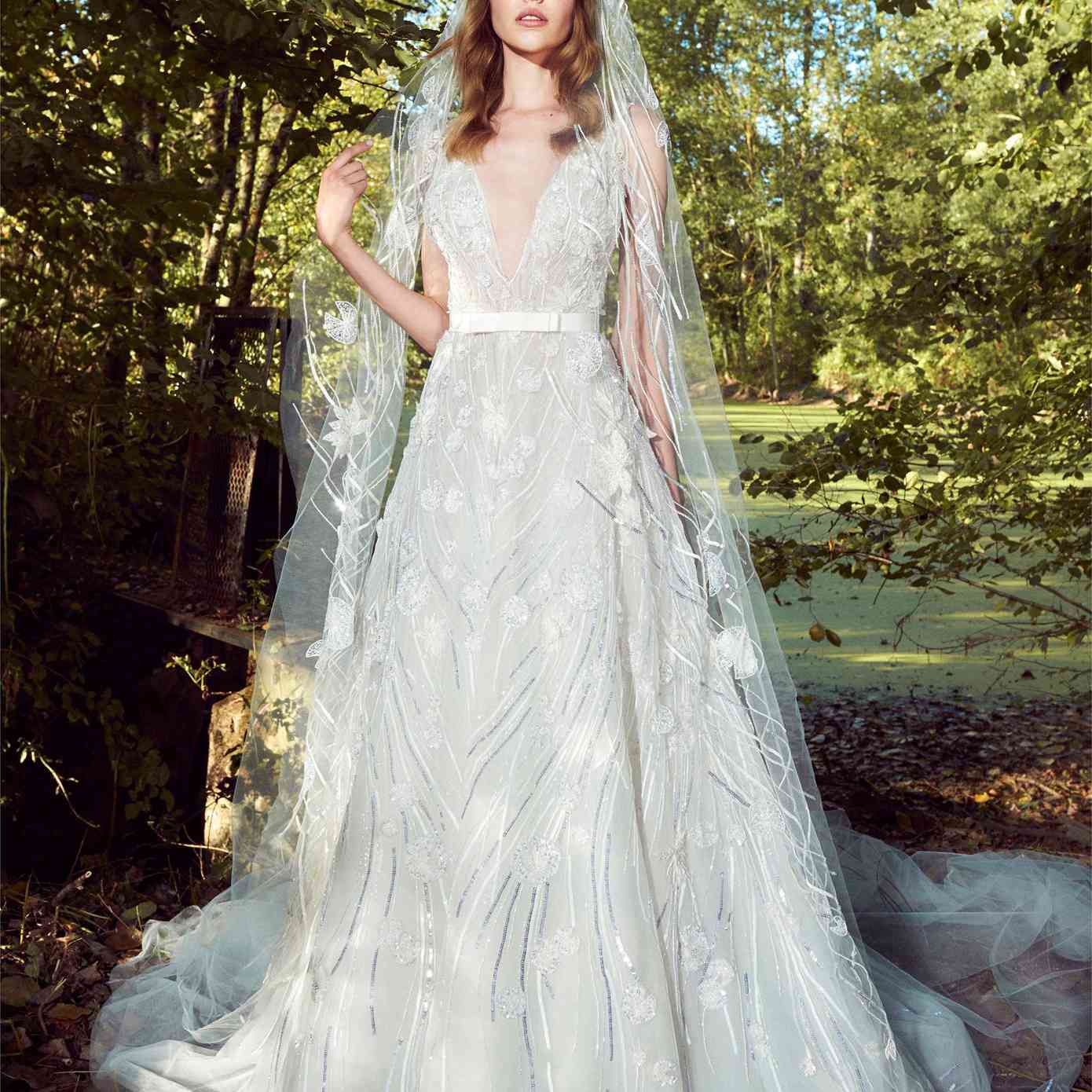 模型在深v领裙袍银和清晰的珠饰和刺绣和一个白色的蝴蝶结腰带和一个匹配的面纱